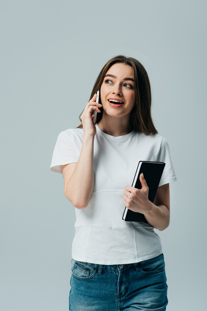 happy beautiful girl in white t-shirt talking on smartphone and holding notebook isolated on grey - Fotó, kép