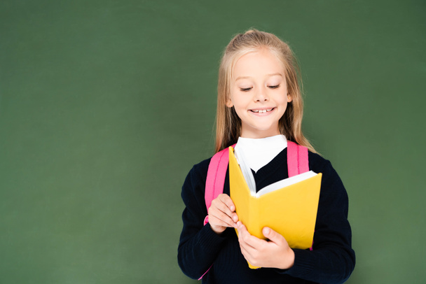 smiling schoolgirl reading book while standing near green chalkboard - Photo, Image
