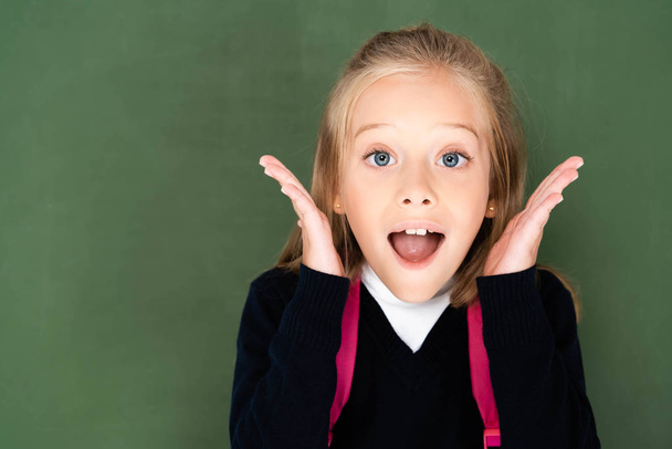 surprised schoolgirl looking at camera while standing near green chalkboard - Photo, Image