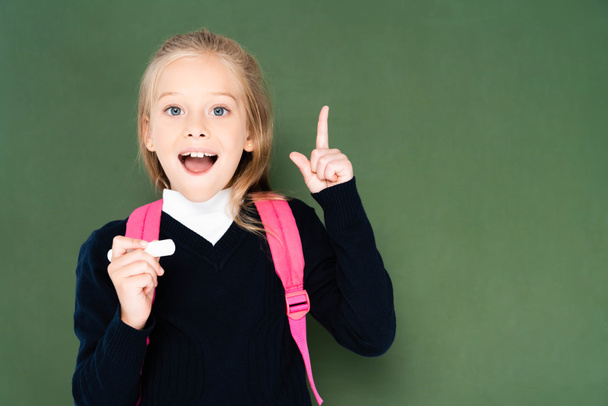 fröhliches Schulmädchen, das ein Stück Kreide in der Hand hält und eine Geste zeigt, während es neben einer grünen Tafel steht - Foto, Bild