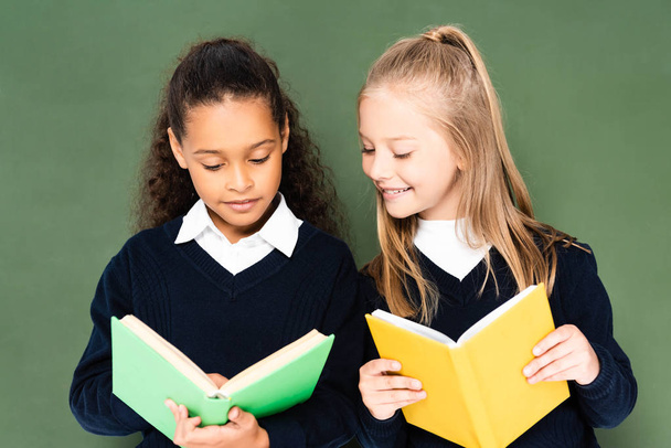 two cheerful multicultural schoolgirls standing reading books together while standing near chalkboard - Photo, Image