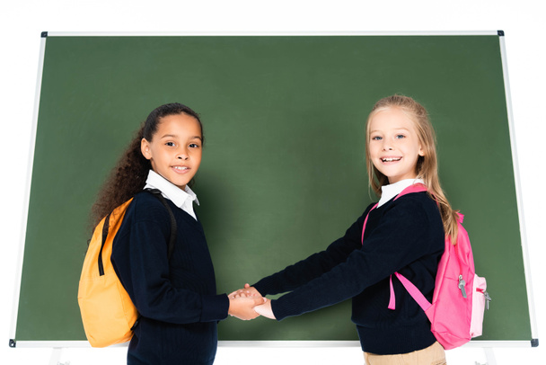 two adorable multicultural schoolgirls holding hands while standing near chalkboard isolated on white - Photo, Image