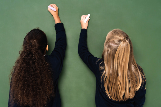 back view of two multicultural schoolgirls writing on chalkboard with chalks - Photo, Image