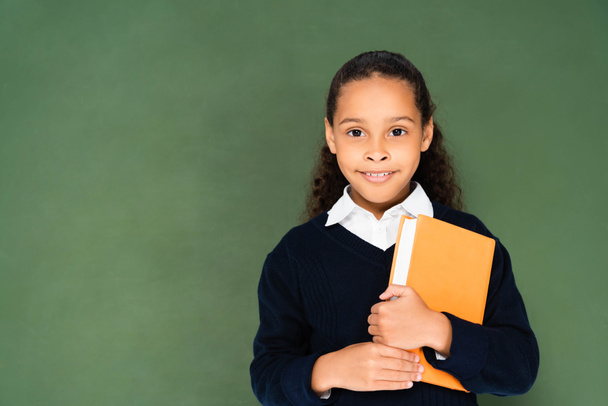 adorable african american schoolgirl holding book and looking at camera while standing near green chalkboard - Foto, imagen