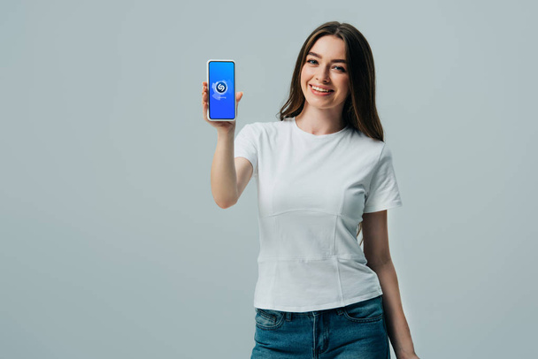 KYIV, UKRAINE - JUNE 6, 2019: happy beautiful girl in white t-shirt showing smartphone with shazam app isolated on grey - Photo, Image