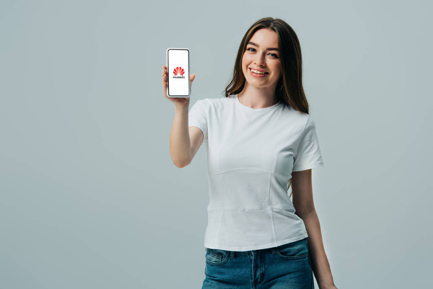 KYIV, UKRAINE - JUNE 6, 2019: happy beautiful girl in white t-shirt showing smartphone with huawei logo isolated on grey - Фото, зображення