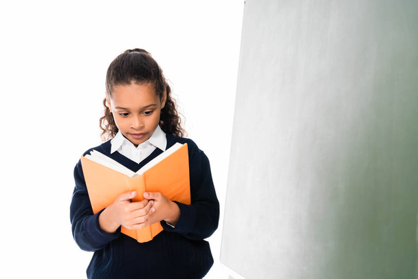 mignon afro-américain écolière lecture livre tout en se tenant près de tableau vert isolé sur blanc
 - Photo, image