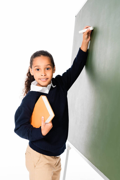 cute african american schoolgirl writing on chalkboard while holding book and looking at camera isolated on white - Фото, изображение