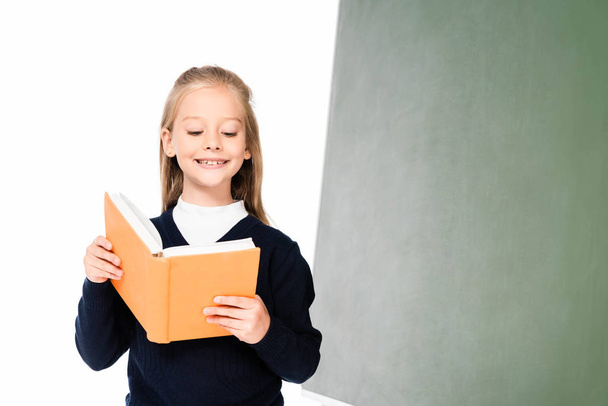 cheerful schoolgirl reading book while standing near chalkboard isolated on white - Photo, Image