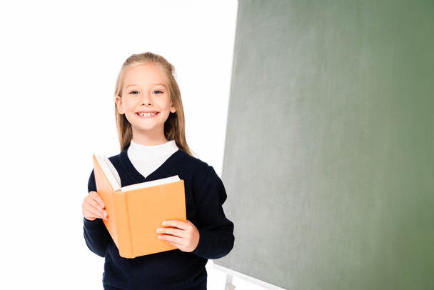 adorable schoolgirl smiling at camera while standing near chalkboard and holding book isolated on white - Photo, image