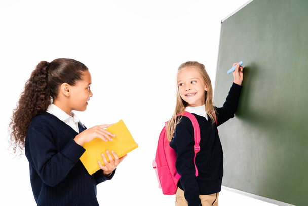 two smiling schoolgirl writing on clipboard and looking at african american classmate holding book isolated on white - Foto, Bild