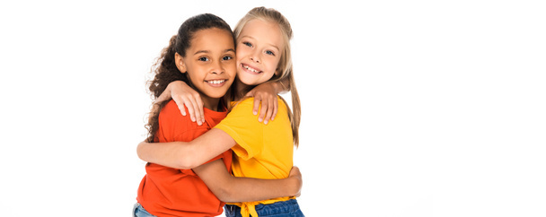 panoramic shot of two happy multicultural friends hugging while looking at camera isolated on white - Foto, afbeelding