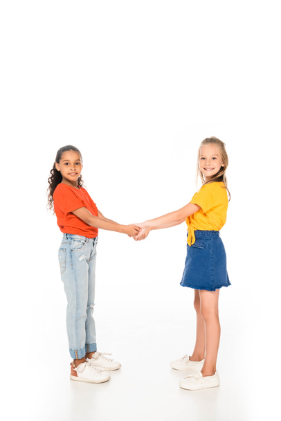 full length view of two cute multicultural friends holding hands and smiling at camera on white background - Фото, зображення