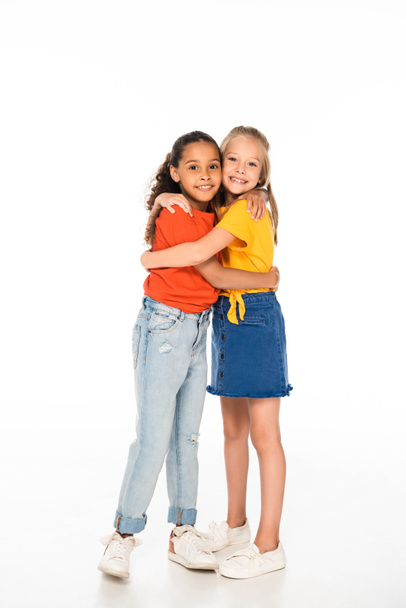 full length view of two happy multicultural friends hugging while smiling at camera on white background - Foto, Bild
