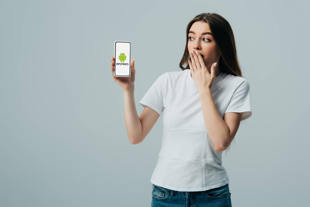KYIV, UKRAINE - JUNE 6, 2019: shocked beautiful girl in white t-shirt showing smartphone with android logo isolated on grey - Foto, Bild