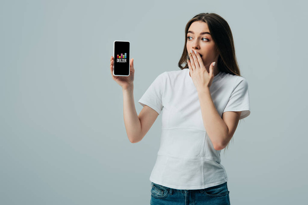 KYIV, UKRAINE - JUNE 6, 2019: shocked beautiful girl in white t-shirt showing smartphone with deezer app isolated on grey - Fotó, kép