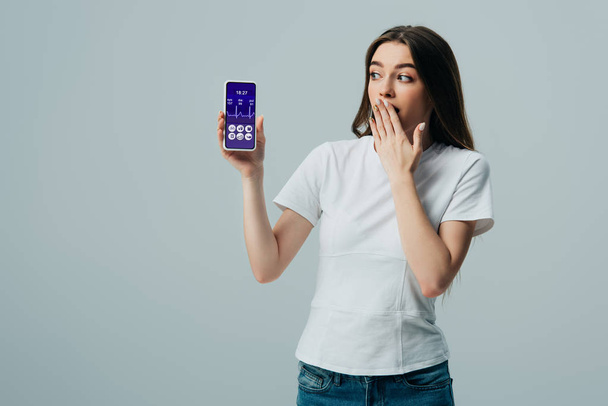shocked beautiful girl in white t-shirt showing smartphone with healthcare app isolated on grey - Photo, Image