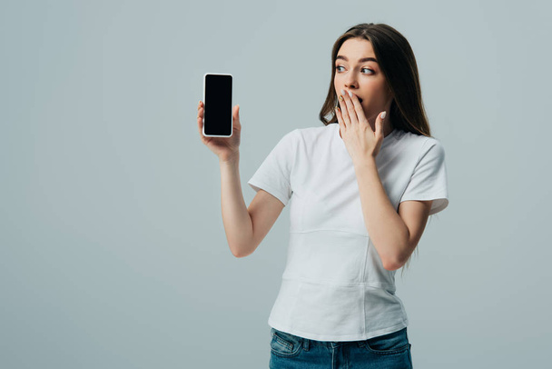 shocked beautiful girl in white t-shirt showing smartphone with blank screen isolated on grey - Valokuva, kuva