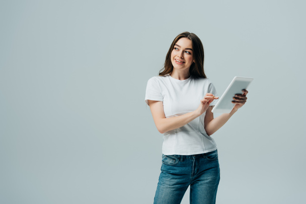happy girl in white t-shirt holding digital tablet and looking away isolated on grey - Фото, изображение
