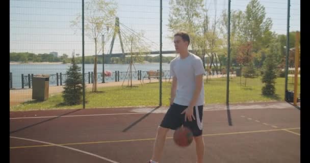 Closeup portrait of young caucasian male basketball player throwing a ball in the hoop on the court outdoors with bridge on the background - Felvétel, videó