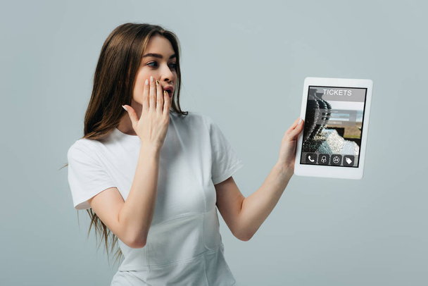 shocked beautiful girl in white t-shirt showing digital tablet with tickets app isolated on grey - Foto, Bild