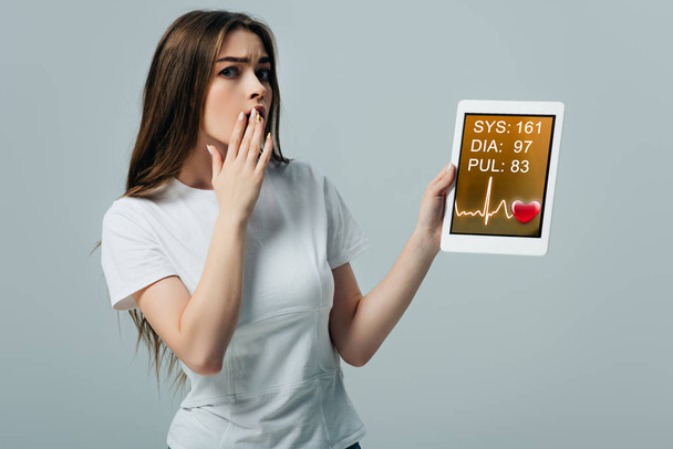 wstrząśnięty piękną dziewczyną w białym t-shirt pokazując cyfrowy tablet z kardiologicznej aplikacji na szarym tle - Zdjęcie, obraz