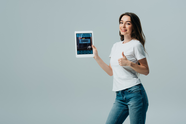 smiling beautiful girl in white t-shirt showing digital tablet with online booking app and showing thumb up isolated on grey - Fotó, kép