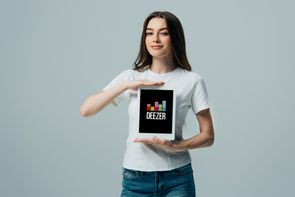 Kiev, Oekraïne-6 juni 2019: glimlachend mooi meisje in wit t-shirt toont digitale tablet met Deezer app geïsoleerd op grijs - Foto, afbeelding