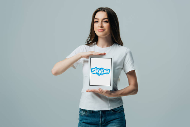 Κίεβο, Ουκρανία-6 Ιουνίου 2019: χαμογελαστός όμορφος κορίτσι σε λευκό μπλουζάκι που δείχνει ψηφιακό δισκίο με εφαρμογή Skype απομονωμένη σε γκρι - Φωτογραφία, εικόνα