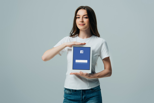 Київ, Україна-6 червня 2019: усміхнена красива дівчина в Білій футболці показує цифровий планшет з додатком Facebook, ізольований на сірому - Фото, зображення