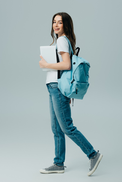 full length view of pretty smiling girl in jeans with blue backpack holding digital tablet isolated on grey - Photo, Image
