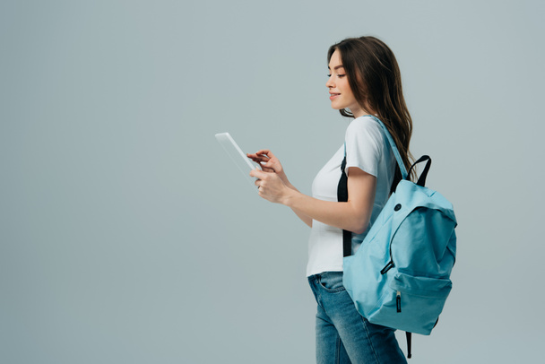 Seitenansicht des hübschen Mädchens mit blauem Rucksack mit digitalem Tablet isoliert auf grau - Foto, Bild