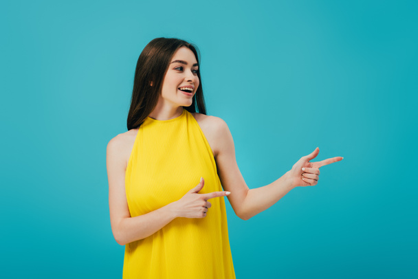 happy beautiful girl in yellow dress pointing with fingers and looking away isolated on turquoise - Foto, Imagem