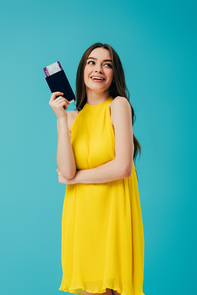 happy brunette woman in yellow dress holding passport with air ticket isolated on turquoise - Zdjęcie, obraz