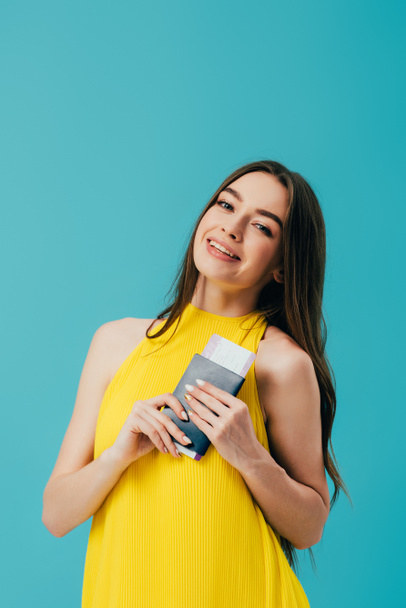happy woman in yellow dress holding passport with air ticket isolated on turquoise - Fotografie, Obrázek