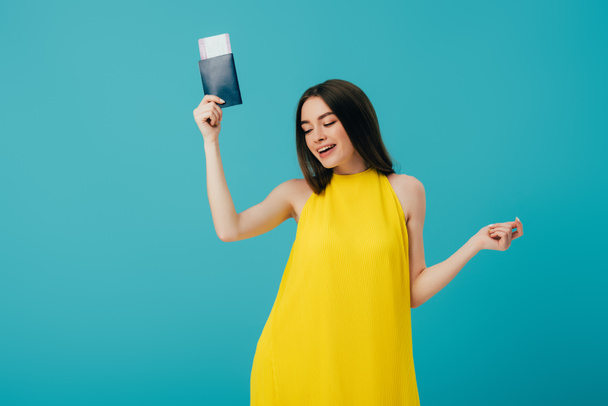 happy brunette woman in yellow dress dancing with passport with air ticket isolated on turquoise - Photo, Image