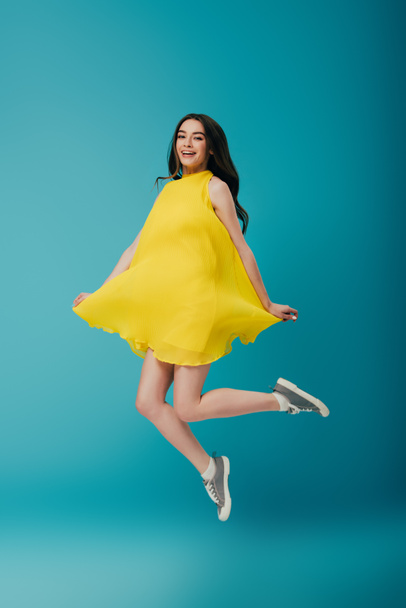 full length view of happy girl in yellow dress jumping on turquoise background - Photo, image
