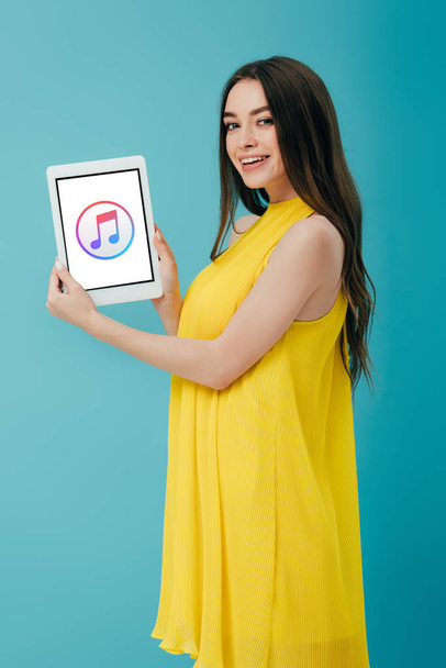 KYIV, UKRAINE - JUNE 6, 2019: smiling beautiful girl in yellow dress showing digital tablet with iTunes app isolated on turquoise - Foto, imagen
