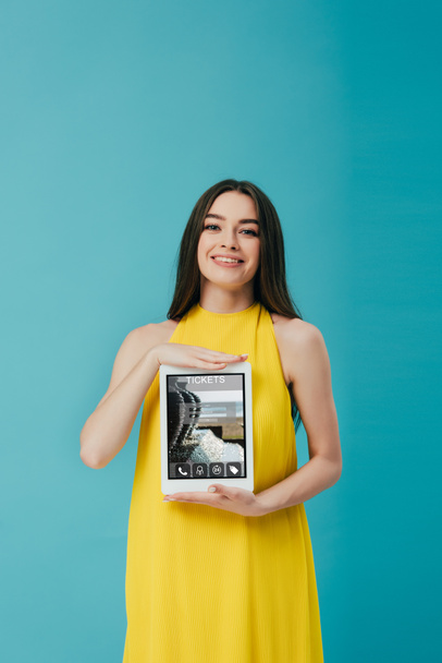 beautiful brunette girl in yellow dress showing digital tablet with tickets app isolated on turquoise - 写真・画像