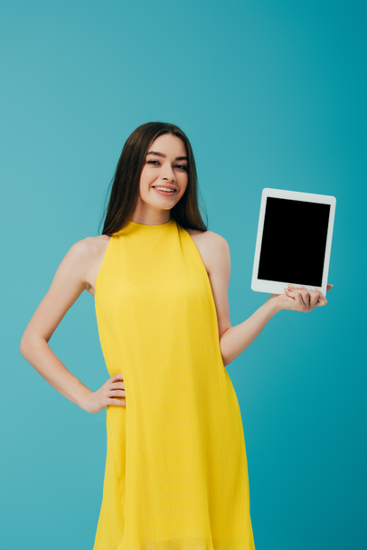 beautiful brunette girl in yellow dress with hand on hip showing digital tablet with blank screen isolated on turquoise - Foto, Imagem