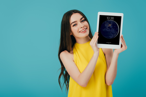 KYIV, UKRAINE - JUNE 6, 2019: happy beautiful girl in yellow dress showing digital tablet with apple lockscreen isolated on turquoise - Photo, image