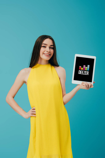 KYIV, UKRAINE - JUNE 6, 2019: smiling girl in yellow dress with hand on hip showing digital tablet with deezer app isolated on turquoise - Photo, Image