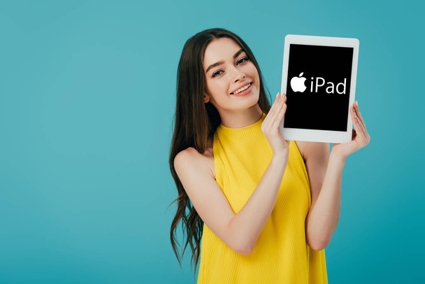 KYIV, UKRAINE - JUNE 6, 2019: happy beautiful girl in yellow dress showing digital tablet with iPad logo isolated on turquoise - Photo, Image