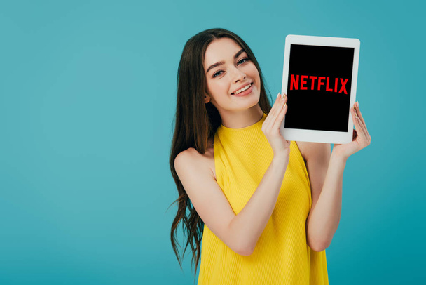 KYIV, UKRAINE - JUNE 6, 2019: happy beautiful girl in yellow dress showing digital tablet with Netflix app isolated on turquoise - Foto, Imagen