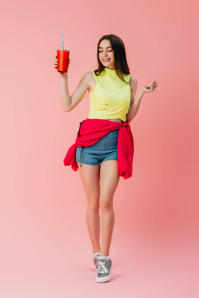повний вигляд красивої щасливої дівчини в яскравому одязі, що тримає соду в пластиковій чашці ізольовані на рожевому
 - Фото, зображення