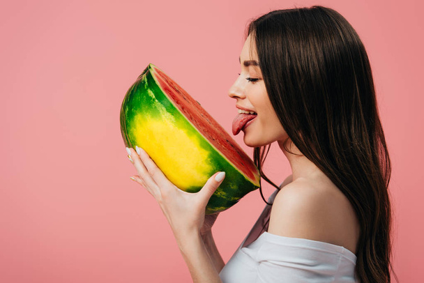 side view of beautiful smiling girl licking ripe delicious watermelon half isolated on pink - 写真・画像
