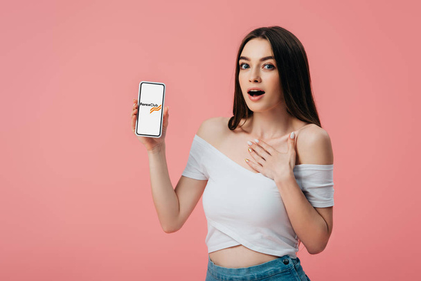 Kyjev, Ukrajina-6. června 2019: krásná šokovaná dívka s smartphone a s klubovou aplikací Forex klubu izolovaná na růžovém - Fotografie, Obrázek