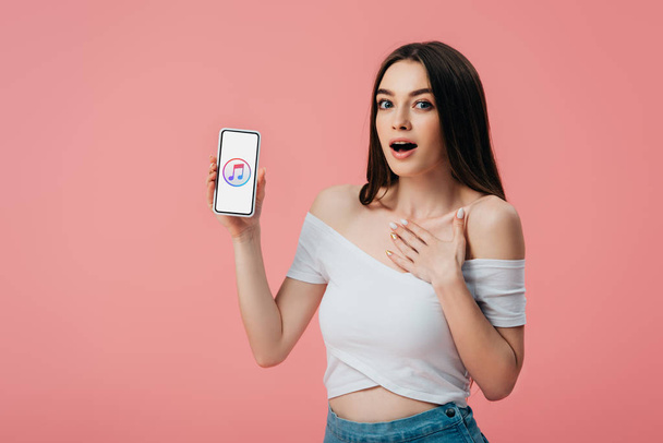 KYIV, UKRAINE - JUNE 6, 2019: beautiful shocked girl holding smartphone with itunes app isolated on pink - Fotografie, Obrázek