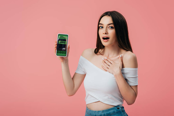 beautiful shocked girl holding smartphone with booking app isolated on pink - Fotó, kép
