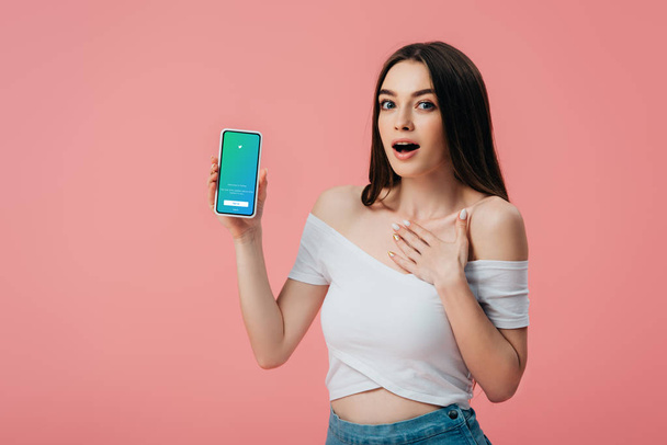 KYIV, UKRAINE - JUNE 6, 2019: beautiful shocked girl holding smartphone with twitter app isolated on pink - Foto, Bild